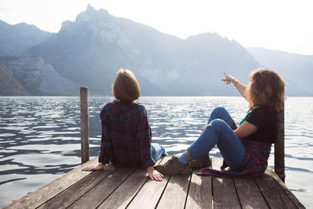Two teenager smiling girls with  sitting on a pier at the lake bank and mountains in the background
の写真素材