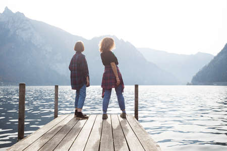 Two teenager smiling girls with  stands on a pier at the lake bank and mountains in the background
の写真素材