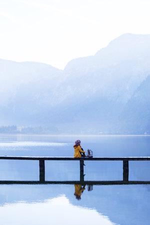 girl tourist in a hat and with a backpack sitting on a wooden bridge on a mountain lake in the early morning. beautiful landscape and reflectionの写真素材