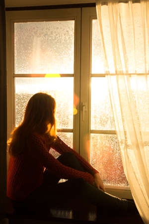 girl sitting on a windowsill and looking at the evening city at the beams of orange sunset sunの写真素材