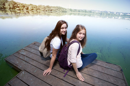 Two teenager girls with backpacks sitting on a pier at the river bank and the city in the background