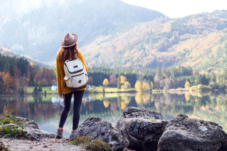 girl in Hat with a backpack stands on the shore of a mountain lake