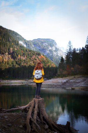 tourist girl with a backpack stands on a huge stump on the shore of a mountain lake