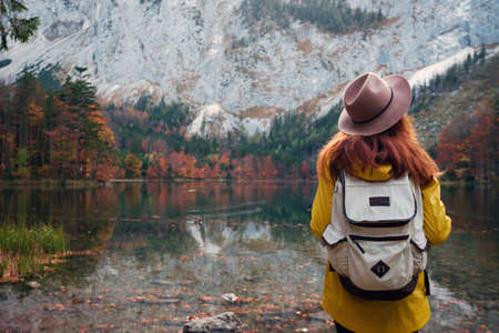 girl on hat with a backpack stands on the shore of a mountain lakeの写真素材