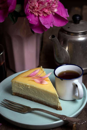 cheesecake on a plate and a teapot, a cup of tea, and a bouquet of peonies nearbyの写真素材
