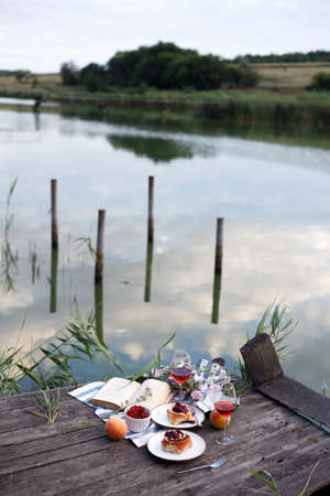 Summer still life-   picnic on a wooden pier. wine, sinabones, peaches, a book with a lake viewの写真素材