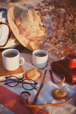 Summer - picnic by the sea. backpack, bouquet, two cups of coffee, cookies, hat, retro camera and book in sunset lightの写真素材