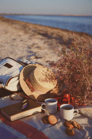 Summer - picnic by the sea. backpack, bouquet, two cups of coffee, cookies, hat, retro camera and book in sunset lightの写真素材