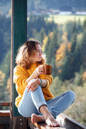 Girl with a cup of coffee sitting on the railing on the balcony. morning in a mountain hutの写真素材
