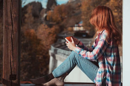 happy smiling girl with a cup of coffee sitting on the railing on the balcony. morning in a mountain hutの写真素材