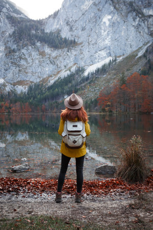 girl on hat with a backpack stands on the shore of a mountain lakeの写真素材