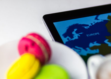 Map of Europe fragment on tablet screen with coffee cup and colorful macaron cakes on white table.