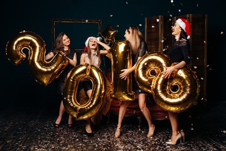 Group of cheerful young women in Christmas hats holding gold colored numbers and throwing confetti at New Year partyの写真素材