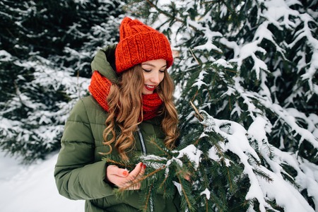 Winter recreation. Young happy girl in winter red clothes walking in winter forest. Happy girl holds snow-covered tree branch in winter forestの写真素材