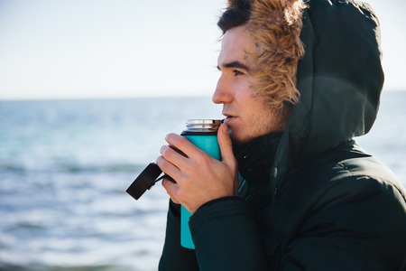 Close-up portrait of young man warming with tea, looking at the sea, dressed in warm jacket with fur hood.の写真素材