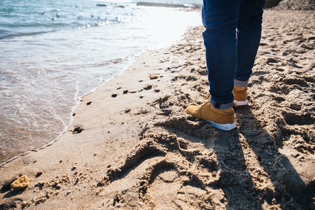 Close-up of men's legs, standing on the sand by the sea. Dressed in jeans, brown boots.の写真素材