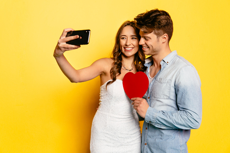 Cheerful woman taking selfie on smartphone with her attractive boyfriend holding red heart. St. Valentine's dayの写真素材