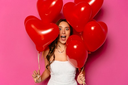 Valentine's day. Wondered young woman with heart shaped air balloons, with mouth wide open, looking at camera. Pink background.の写真素材
