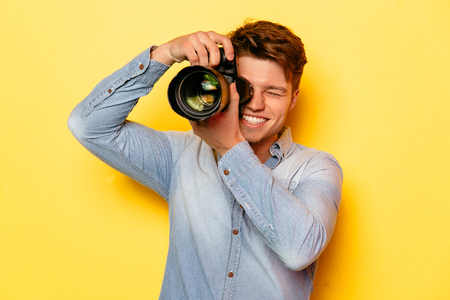 Handsome young photographer with professional camera, taking a photo. On yellow background. Dressed in jeans shirt.の写真素材