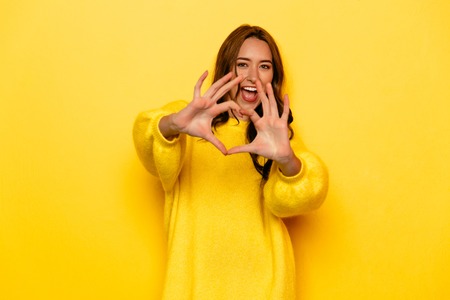 Happy gorgeous young girl in yellow sweater showing heart with two hands, love sign. Isolated over yellow background.の写真素材