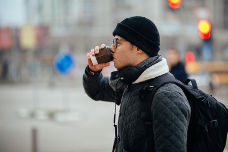 Attractive guy in eyeglasses drinking coffee, outdoors. Dressed in warm jacket and hat, with headphones and school bag. Side view.の写真素材