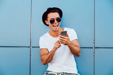 Cheerful handsome man in sunglasses and hat typing a message on smartphone, having a chat, smiling widely. Outdoors.の写真素材