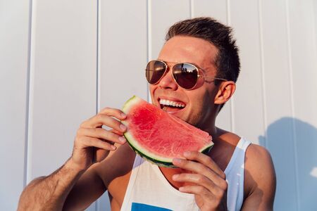 Handsome young man in sunglasses eating a watermelon, enjoying the summer days, outdoors. Dressed in sleeveless t-shirt.の写真素材