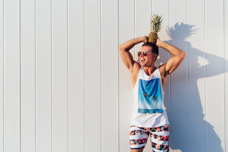 Outdoor photo of cheerful muscular man in sunglasses holding a pineapple on his head, looking away...