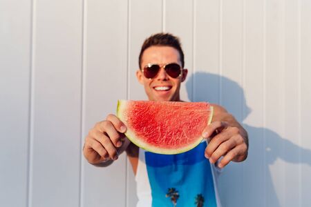 Cheerful smiling man in sunglasses holds a piece of watermelon, looking at camera, outdoors. Dressed in sleeveless t-shirt.の写真素材