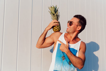 Handsome funny man in sunglasses holds a pineapple on his shoulder and pointing on it, standing against the white wall with copy space. Outdoors.の写真素材