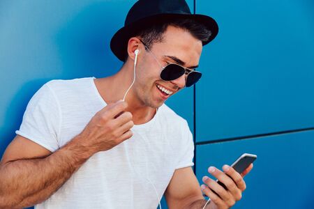 Attractive smiling man in sunglasses looking at smartphone screen, watching a movie in earphones. Wearing stylish clothes. Standing near the wall, outdoors.の写真素材