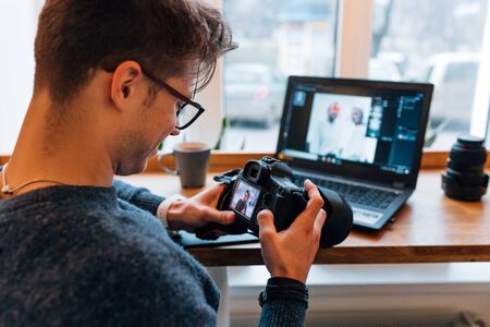 Creative photographer holding professional camera, sitting at cafe with laptop, editing photos. Dressed in sweater, in eyeglasses. Close-up.の写真素材
