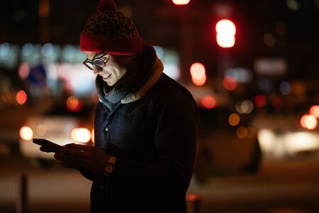Cheerful young man in eyeglasses using his smartphone on background illumination light in night city. Dressed in warm hat and jacket. Outdoors.の写真素材