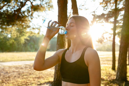 Side view of young sportive woman drinking a water from bottle after running in the park, outdoors....