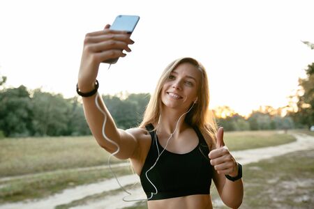 Happy amazing girl in headphones, takes a selfie on mobile phone, showing a thumb up, wearing sport tank top. Outdoors.の写真素材