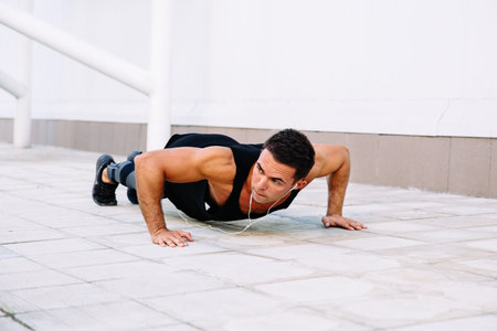 Attractive muscular guy doing push-ups exercises, during workout outdoors, listening to music in...