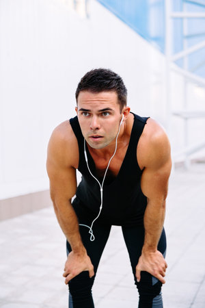 Portrait of tired sportsman, resting after running, keeping hands on his knees, listening to music...