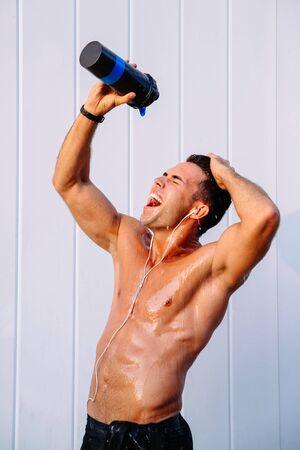 Side view of happy muscular guy pouring out water from bottle on his body, sweating after training hard. Outdoors.の写真素材