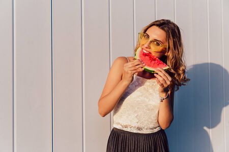 Curly young woman in sunglasses eating a watermelon, enjoying the sunny days, standing against the white wall, outdoors. Dressed in summer blouse and skirt.の写真素材