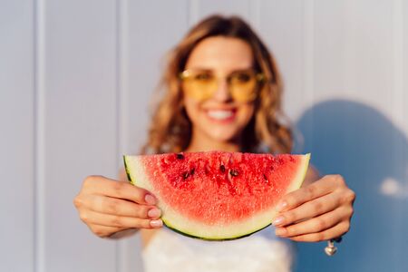 Blurred photo of charming joyful girl holding a piece of juicy watermelon with seeds, standing outdoors.の写真素材