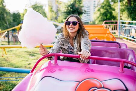 Beautiful cheerful girl riding a roller coaster, holding a cotton candy, wearing sunglasses. Dressed in stylish jacket. Outdoors.の写真素材