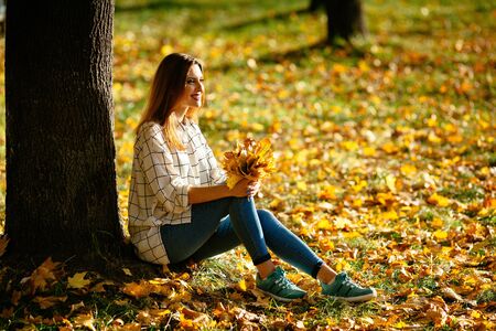 Pretty girl posing while sitting under the tree with a bunch of leaves in her hands.の写真素材