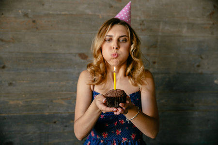 Beautiful young woman blowing out candle on birthday cupcake. Festive hat, curly hair. Holidays...