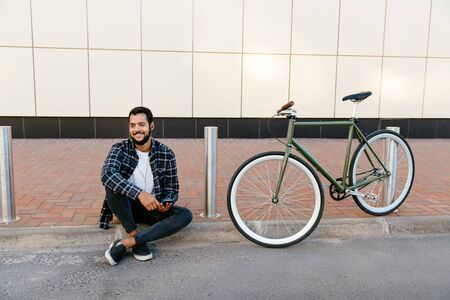 Outdoor photo of stylish attractive man sitting near the bike, listening to music in earphones, enjoying the leisure outside.の写真素材