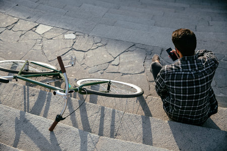 Top view on trendy man using a mobile phone, while resting on steps, sitting near the bike outdoors....