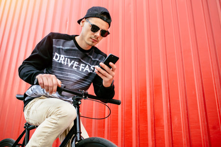 Attractive thoughtful hipster man using a mobile phone, during sitting on bike, resting. Outdoors....