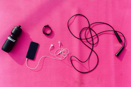 View from top on mobile phone with earphones, jump rope and bottle of water against the pink...