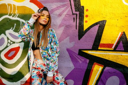 Amazing young model in sunglasses, posing near the wall with graffiti. Dressed in fashionable suit, singlet and cap. Outdoors.の写真素材