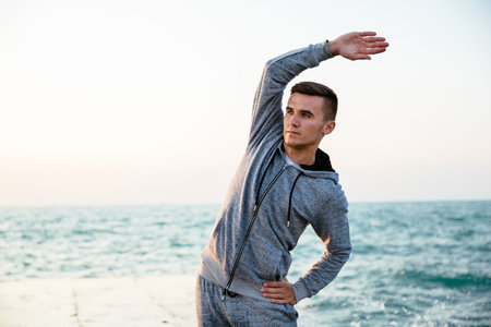 Outdoor photo of young sportsman with serious face in sportswear doing exercises for body, on...