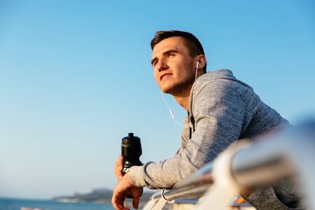 Young handsome sportsman holding a bottle of water, resting after workout, standing on quay and looking at the sea. Sport concept.の写真素材